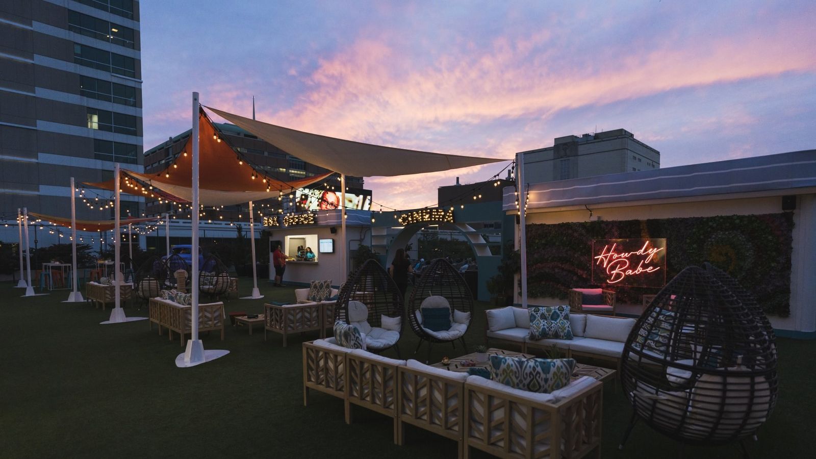 A rooftop lounge with sofas and hanging chairs under shade sails at dusk, with city buildings behind.