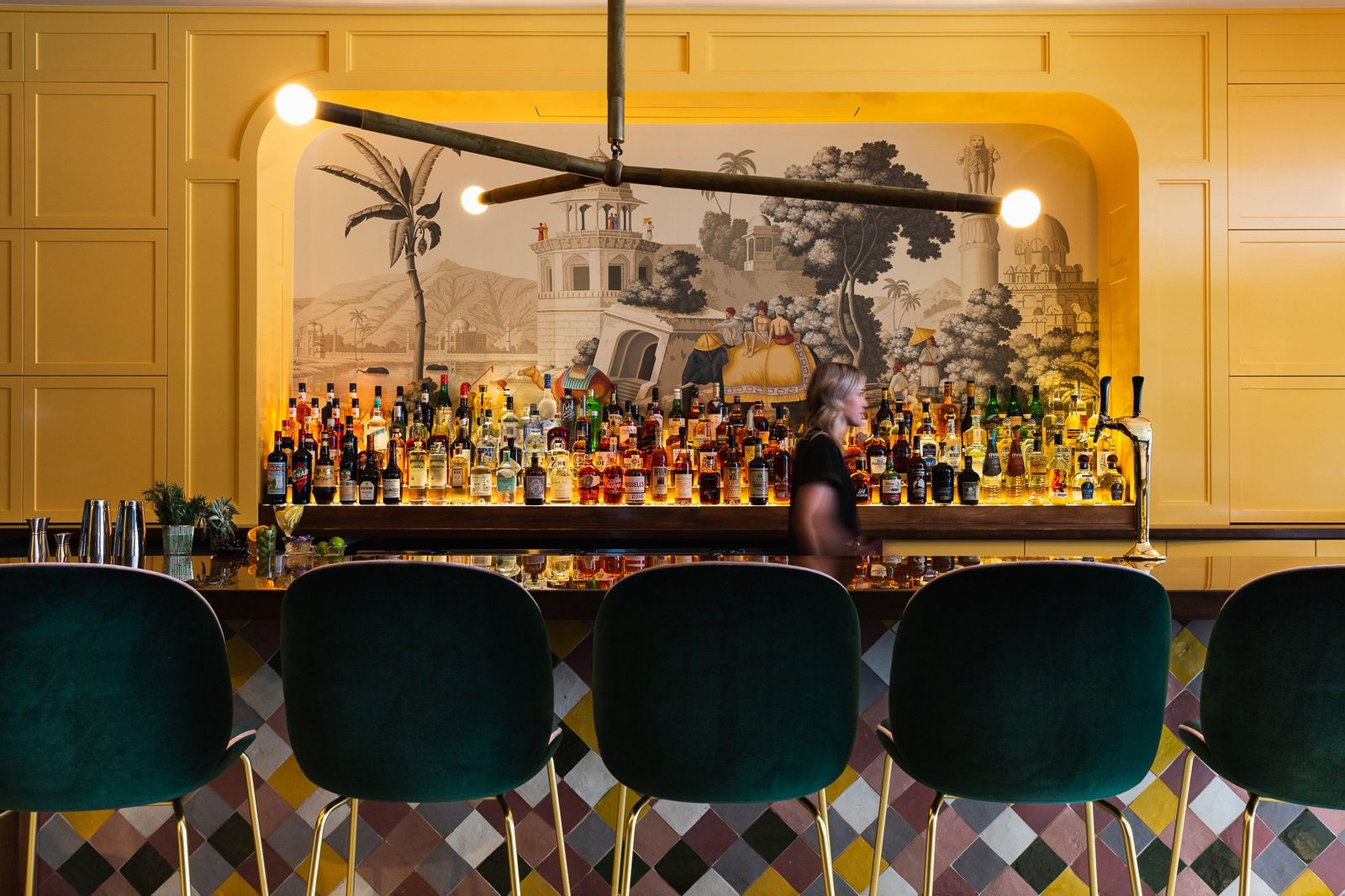 A bar with green velvet stools in front of a lit shelf of bottles and a painted landscape mural.