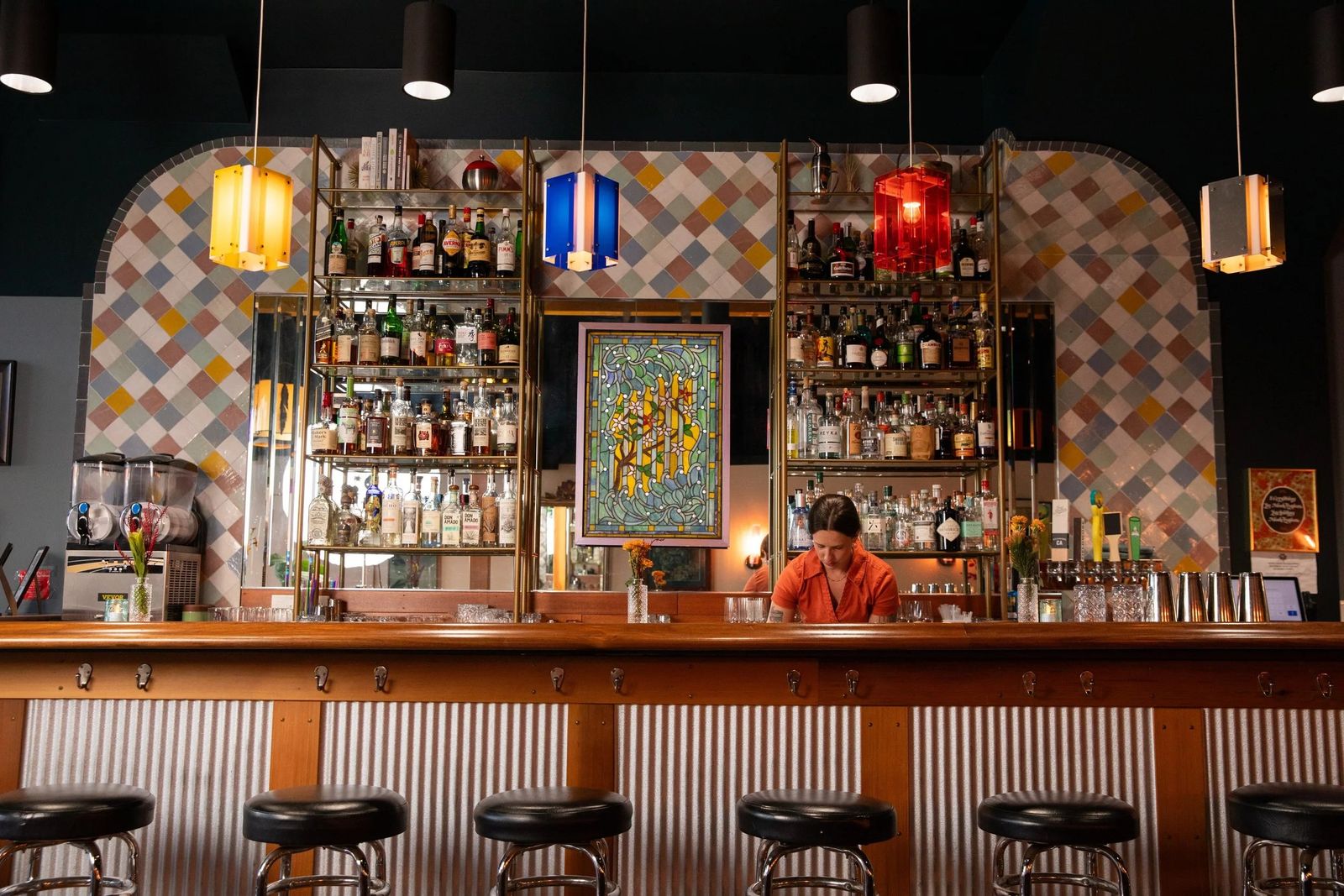 A bar with a tiled back wall, lit shelves of bottles, hanging colored lamps and a bartender behind the counter.