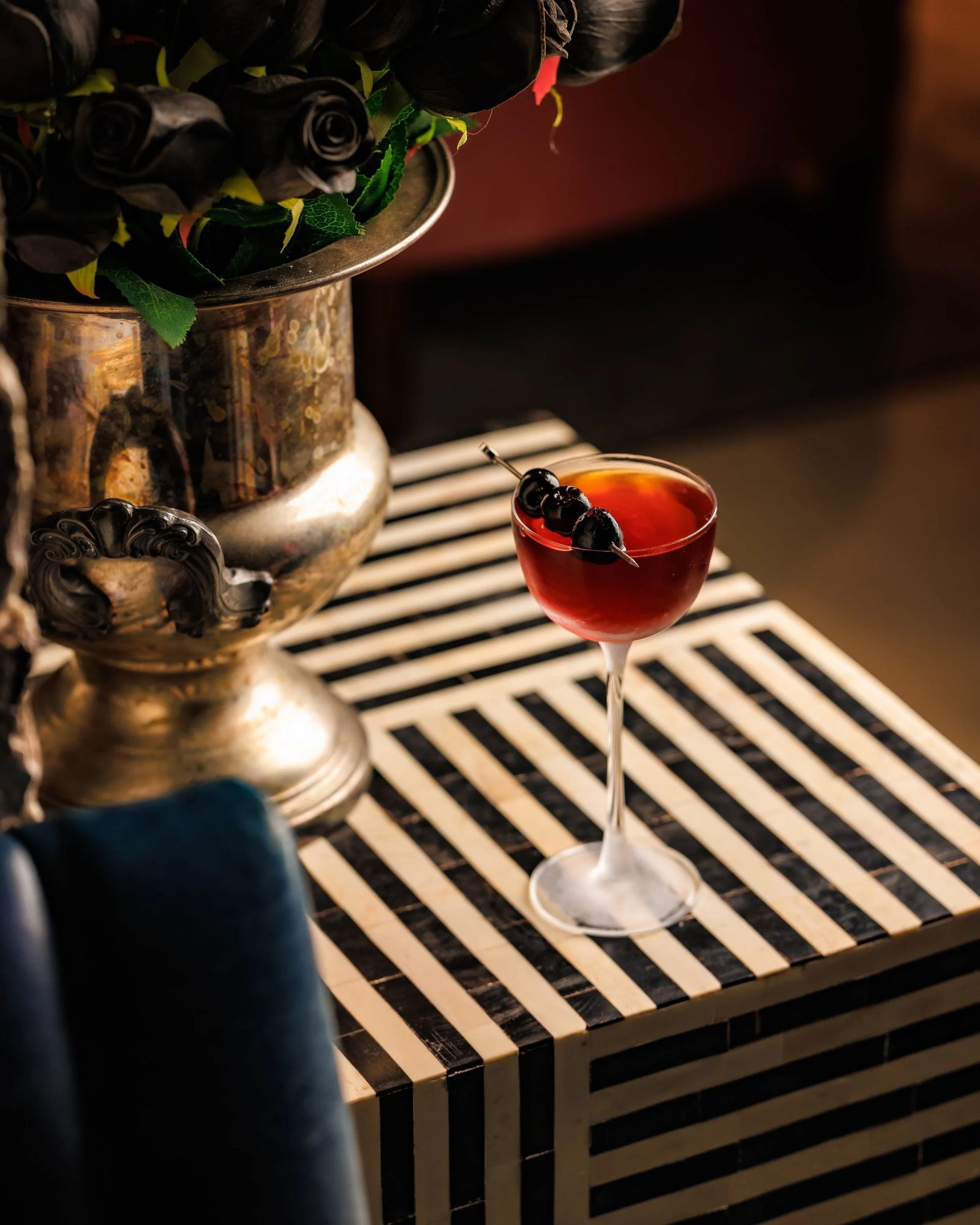 A cocktail with cherries on a black-and-white striped table beside a gold urn of dark roses.