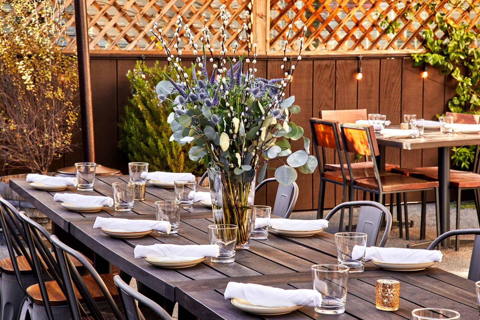 Long wooden table set with glassware and a flower arrangement on a trellised outdoor patio