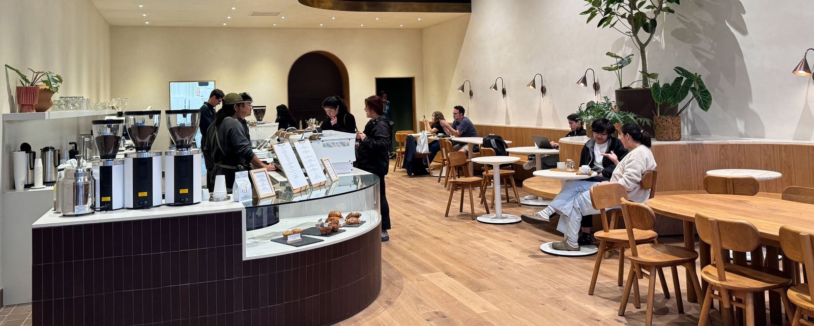 A cafe with a curved pastry counter and espresso machines, with wooden tables and customers in the background.