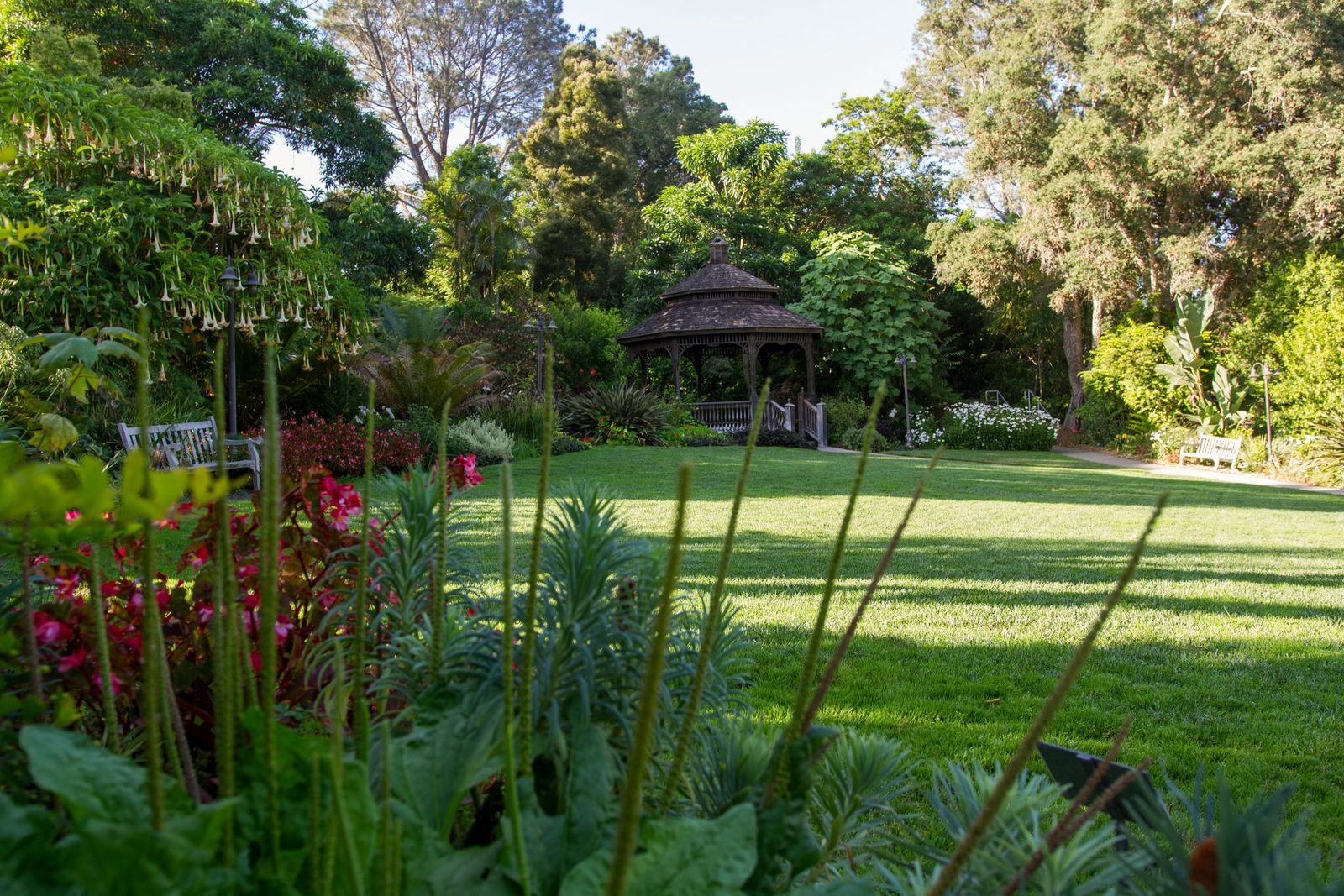 A green lawn bordered by flower beds and trees with a small gazebo in the distance.