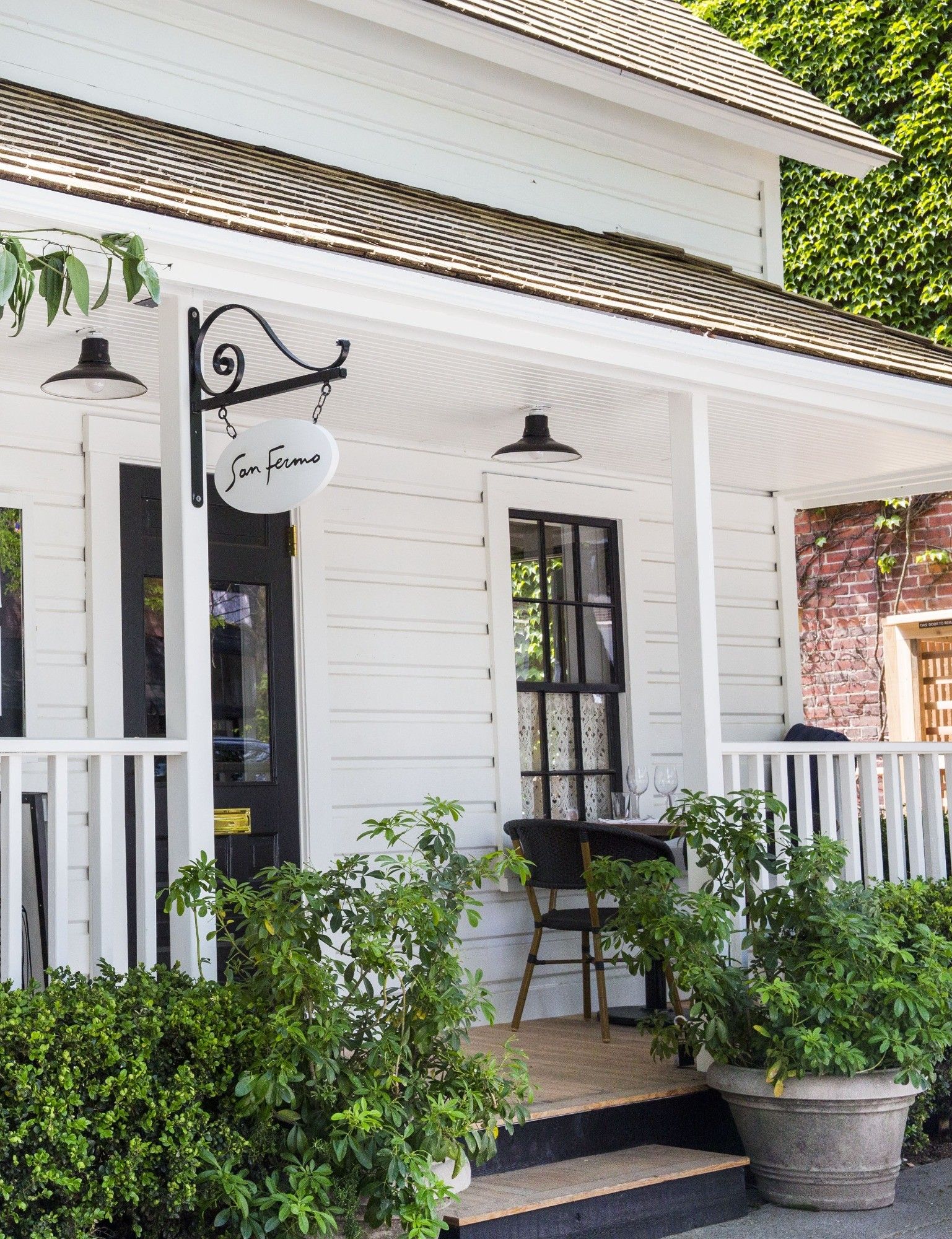 The white clapboard front of the restaurant with a covered porch, a hanging sign and potted plants.