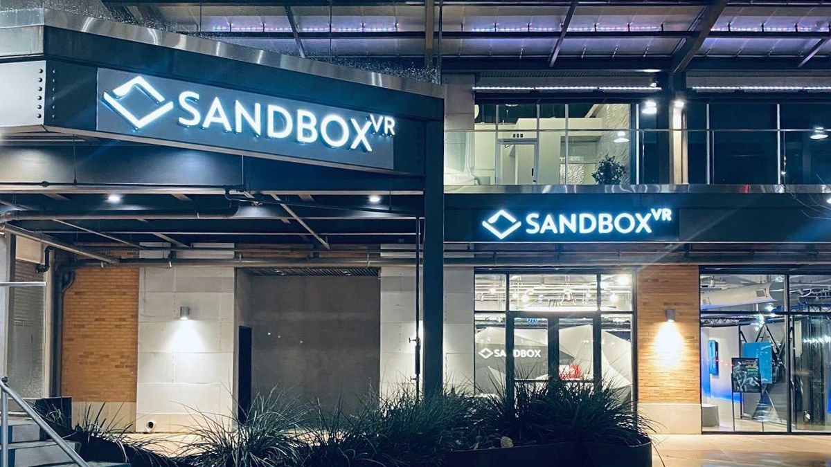 A storefront at night with a large lit Sandbox VR sign above glass windows and a wood-paneled wall.