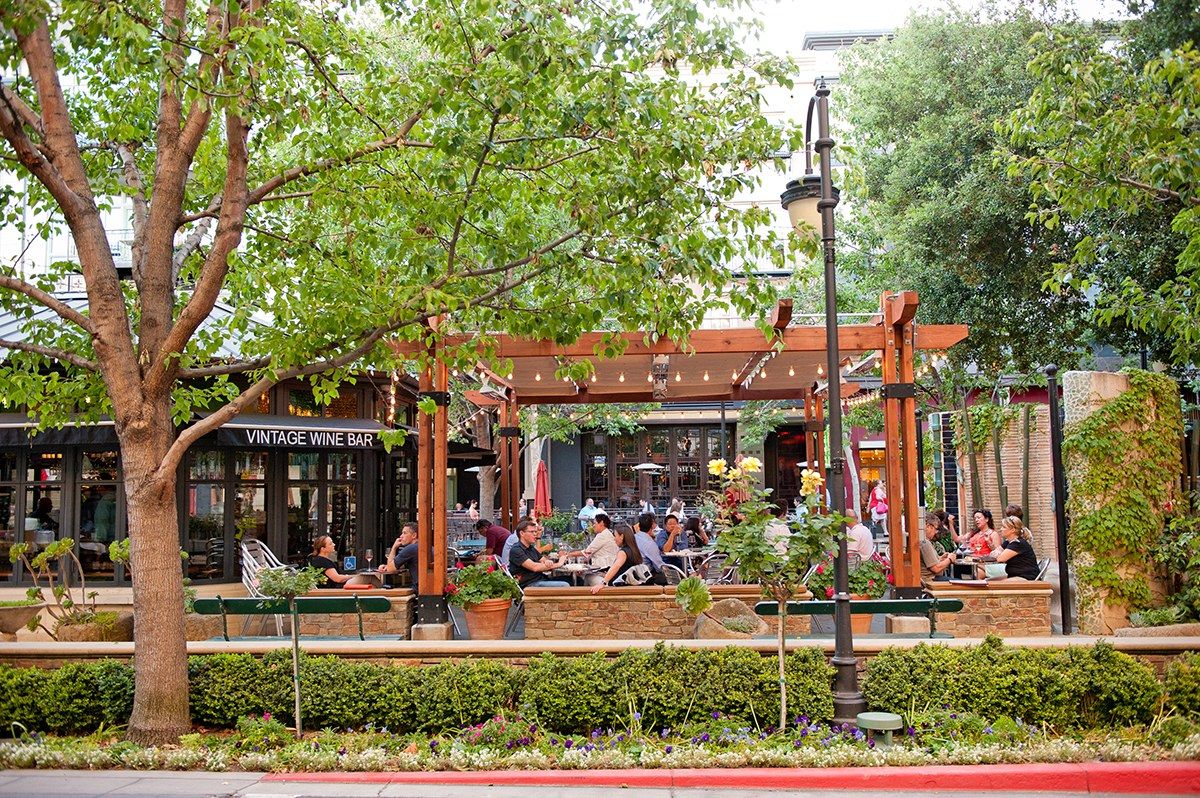 Tree-lined outdoor plaza with restaurant patios and diners beneath a trellis