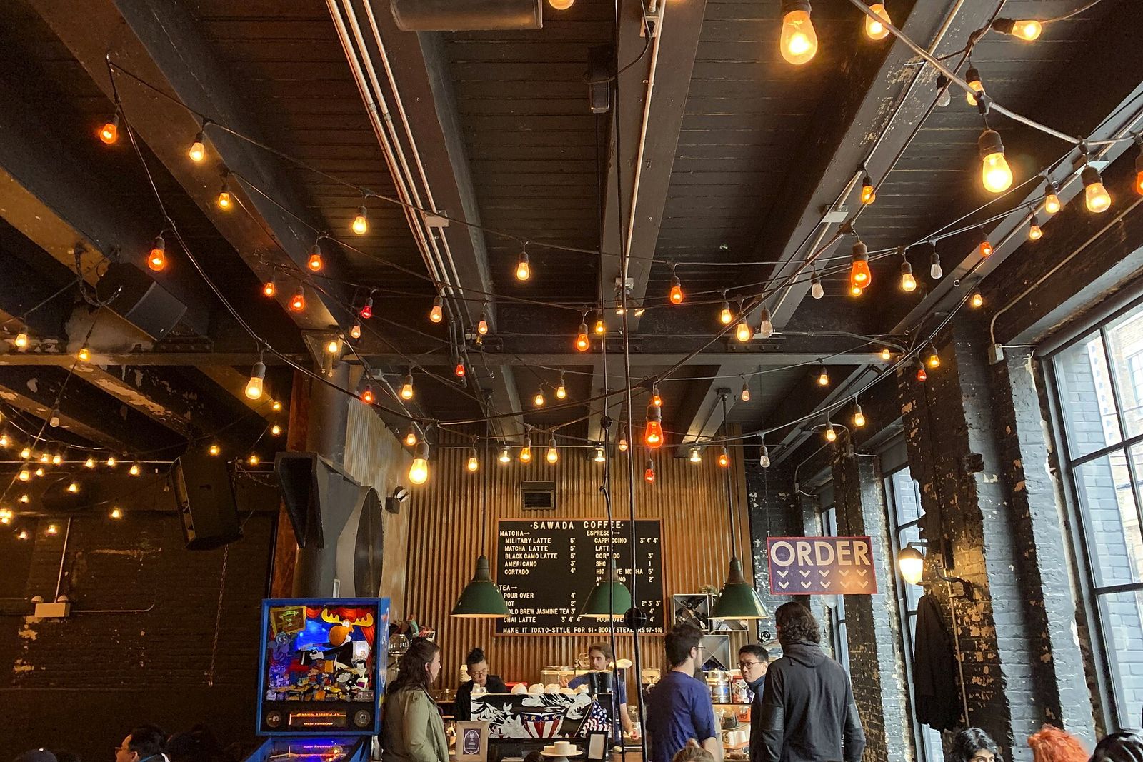 A cafe interior with string lights across the ceiling, a menu board, an arcade game and customers at the counter.