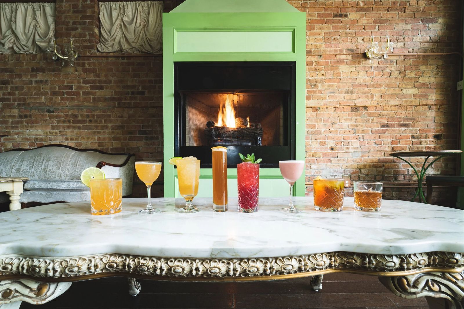 A marble table with a row of cocktails in front of a lit fireplace and a mint-green wall.