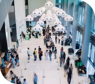 A bright atrium lobby seen from above, with visitors below and rows of white rounded lights hanging from the ceiling.