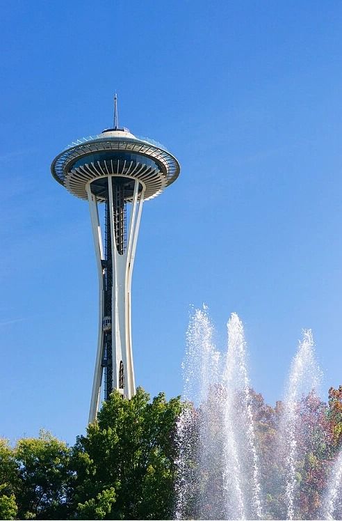 The Space Needle's saucer-shaped top rising above green trees and tall fountain jets under a clear blue sky.