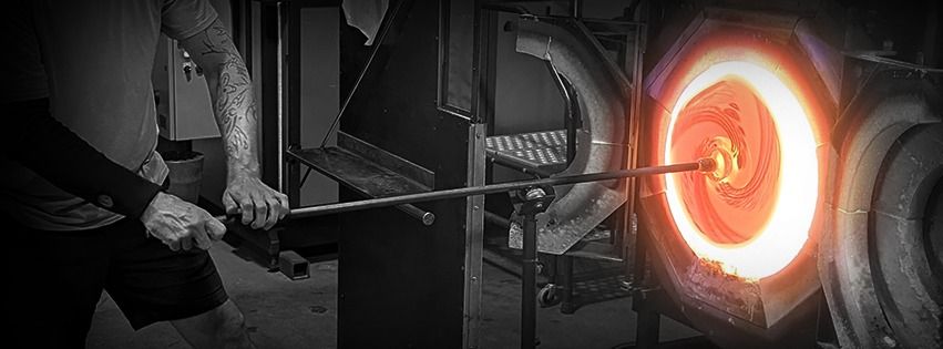 A glassblower holding a long rod to a glowing orange furnace opening, in a black-and-white photo with the furnace in color.