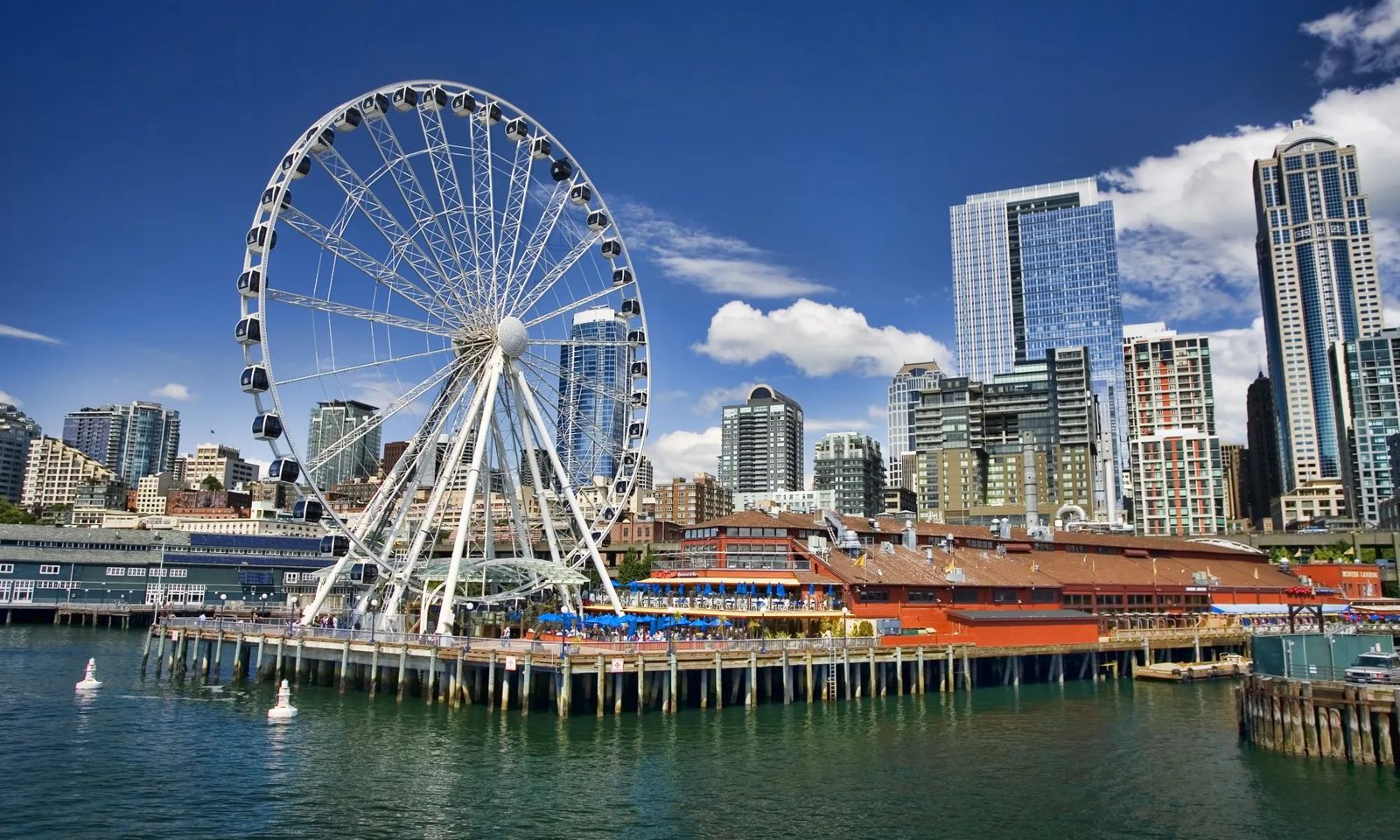 A white Ferris wheel on a pier over the water with downtown towers behind it under a blue sky.