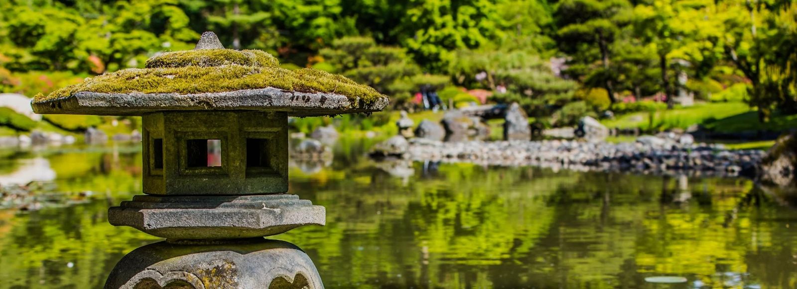A stone lantern beside a still pond, with moss-covered rocks and green trees around it.