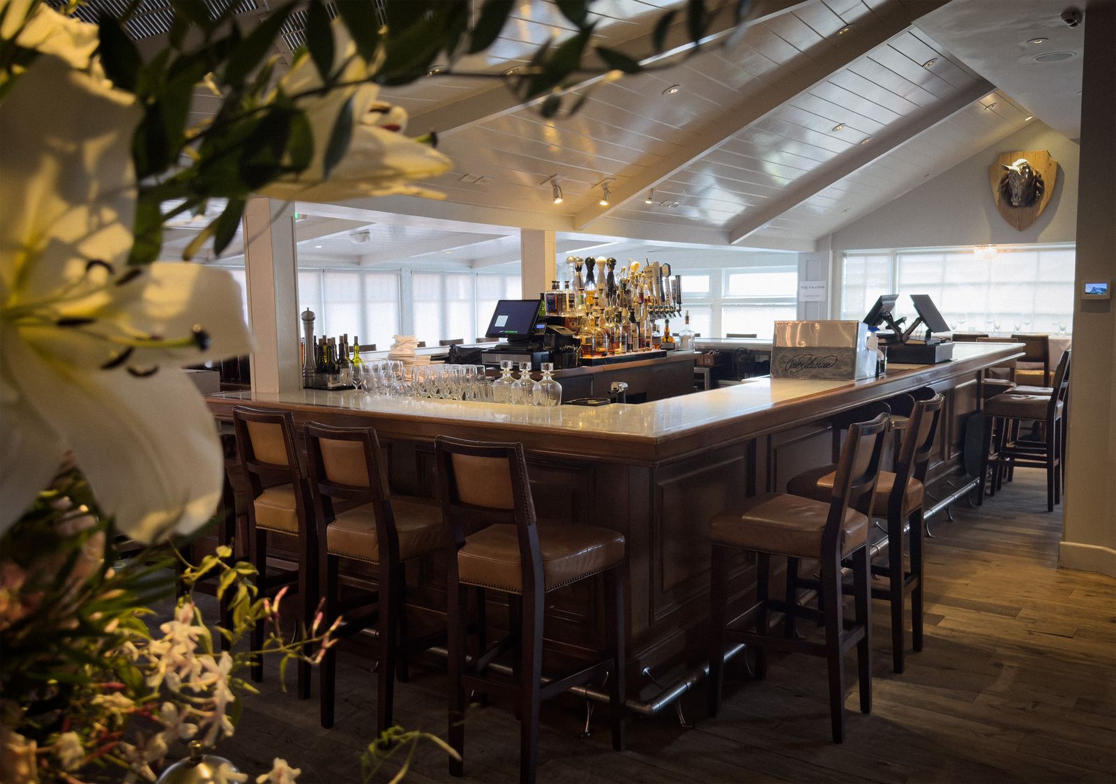 A wood bar with high stools and shelves of bottles in a bright room with flowers in the foreground.