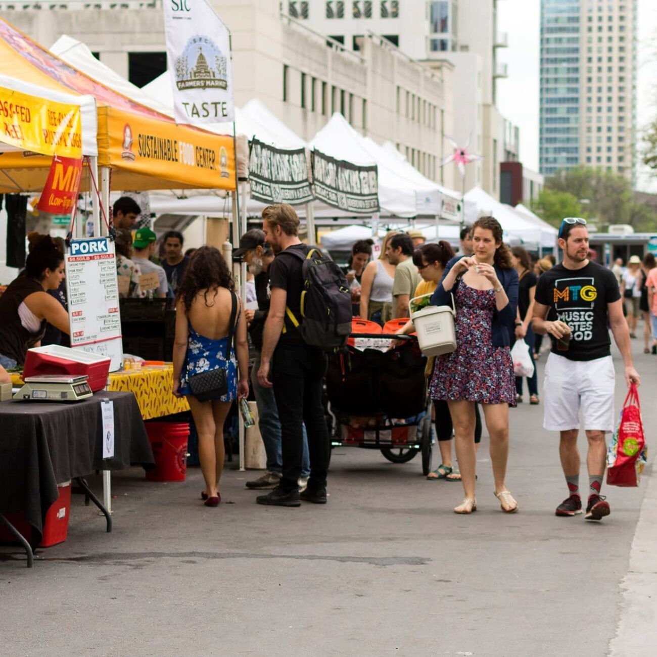 A busy farmers market street with white tents, signs and shoppers, with city buildings behind.