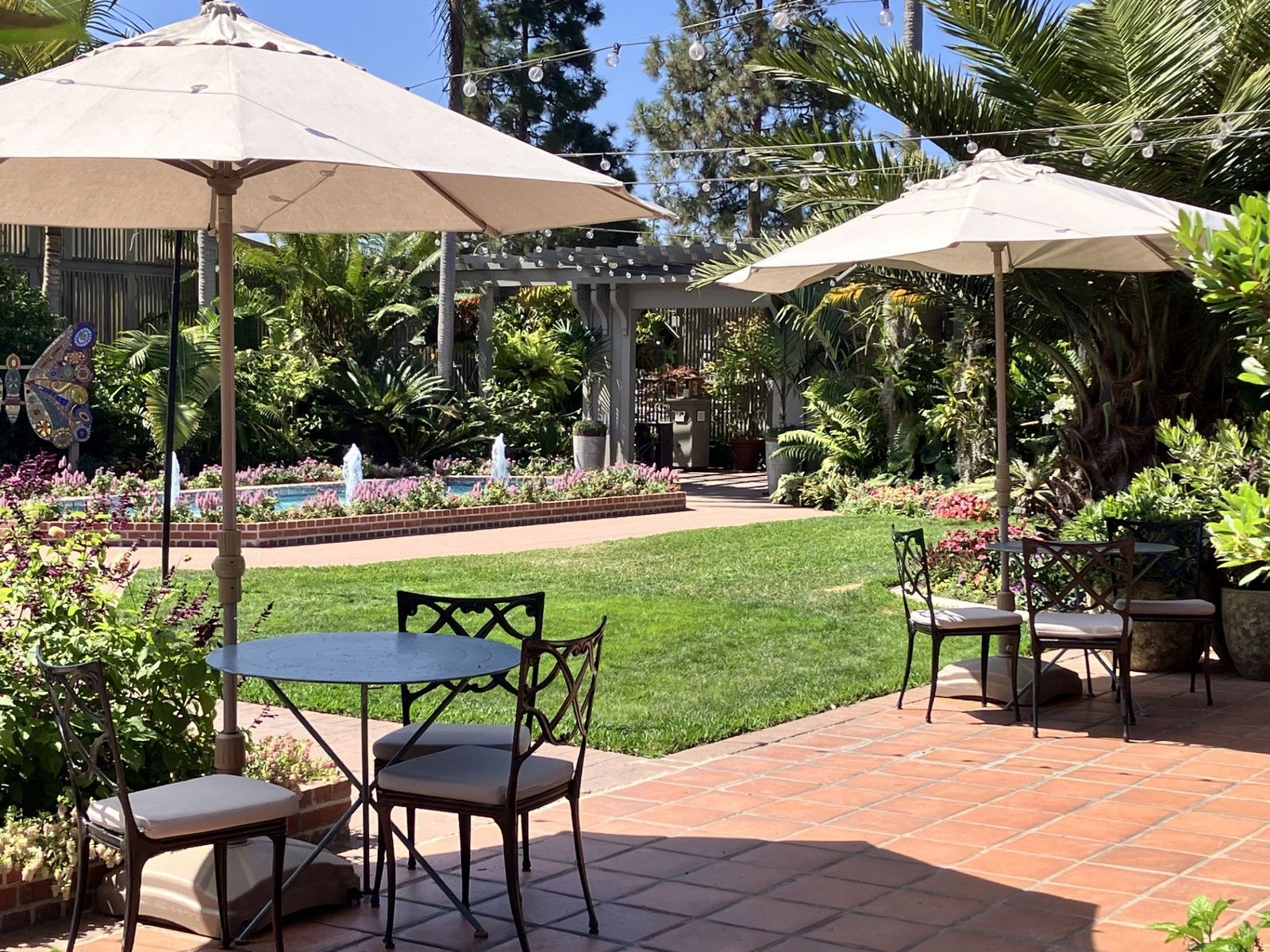 Patio tables with umbrellas on a brick terrace beside a lawn and flower beds.
