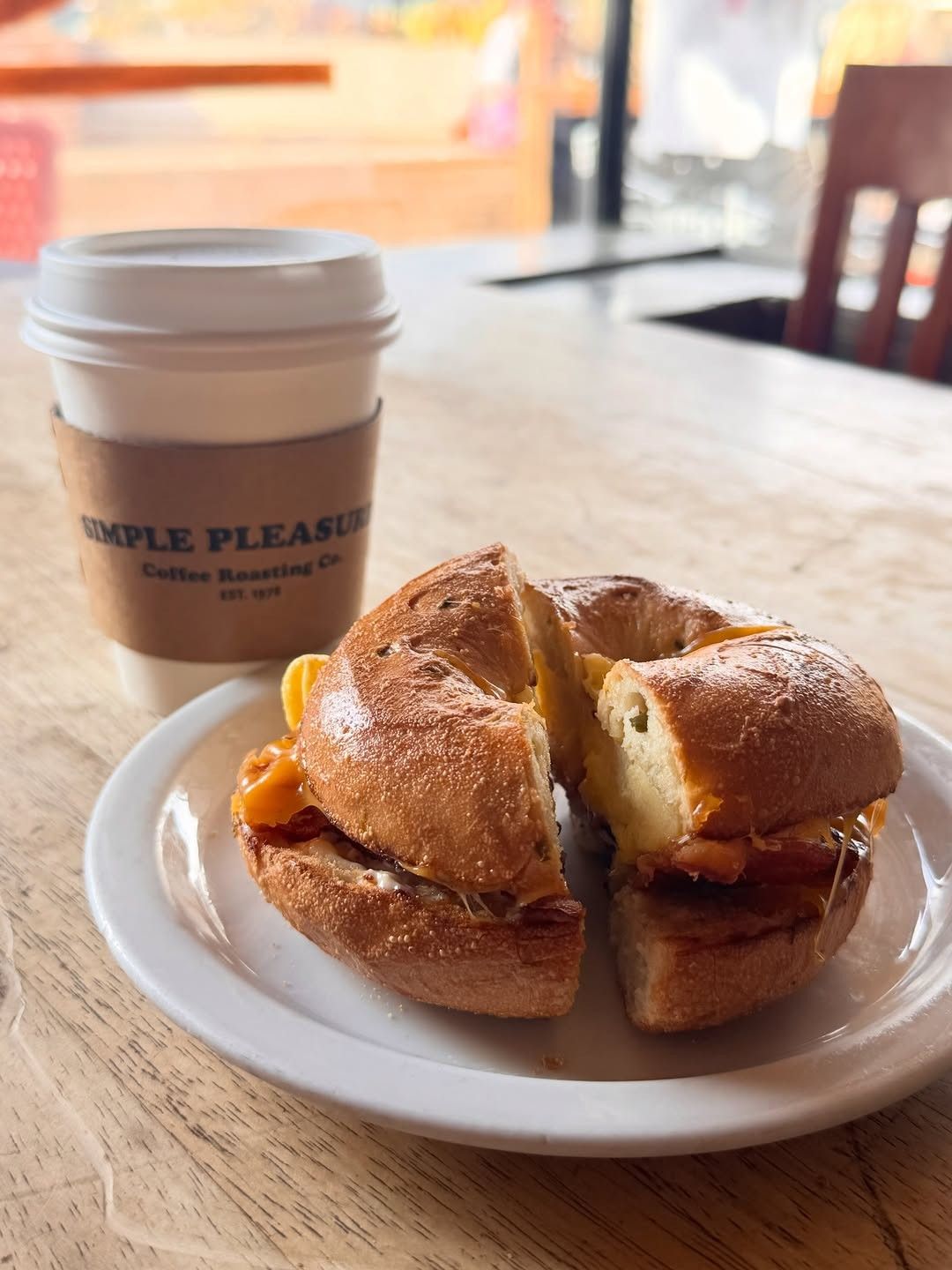 A bagel sandwich on a white plate beside a coffee cup with a lid, on a wooden table.