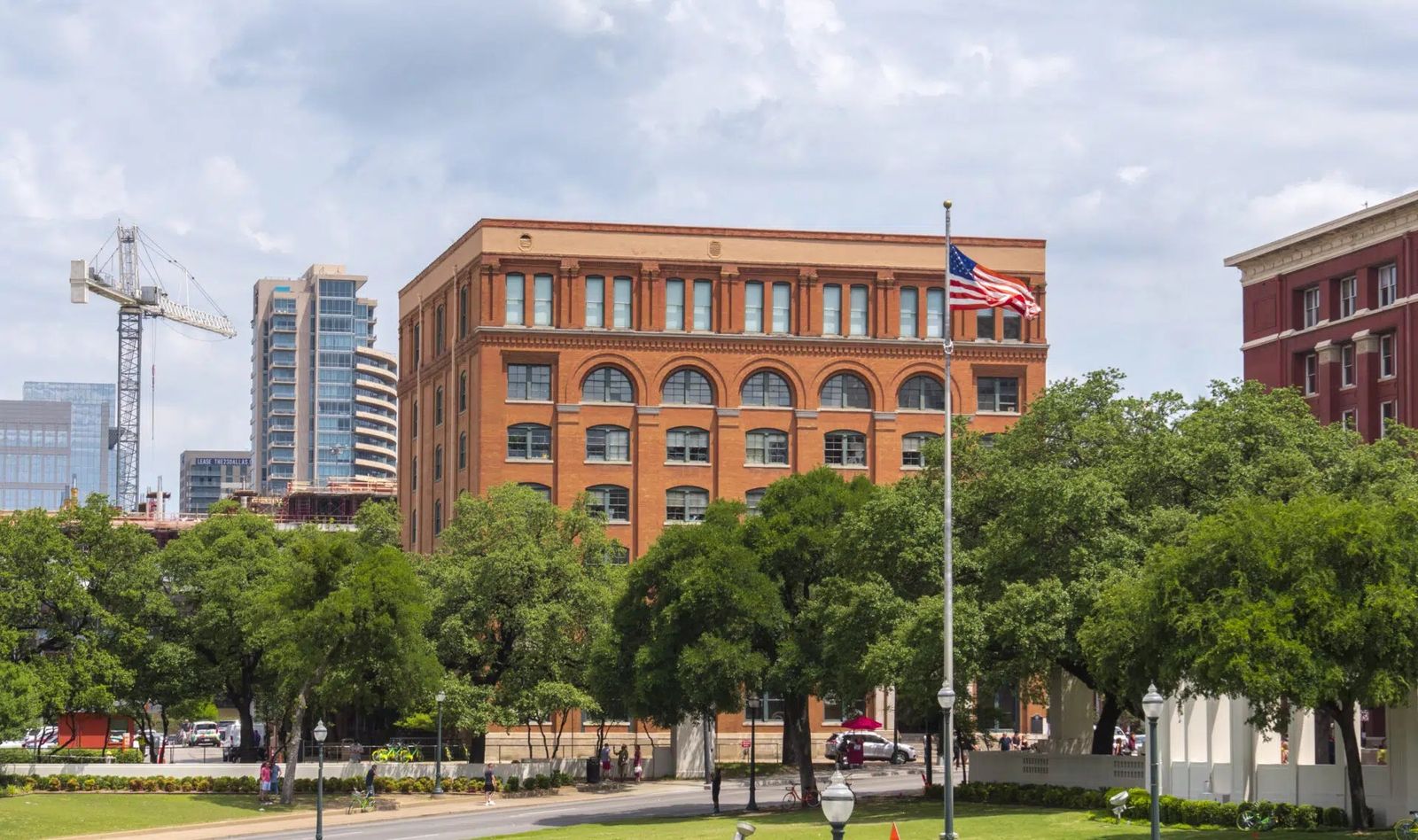 The red brick former book depository building with a flag, seen over trees and a lawn.
