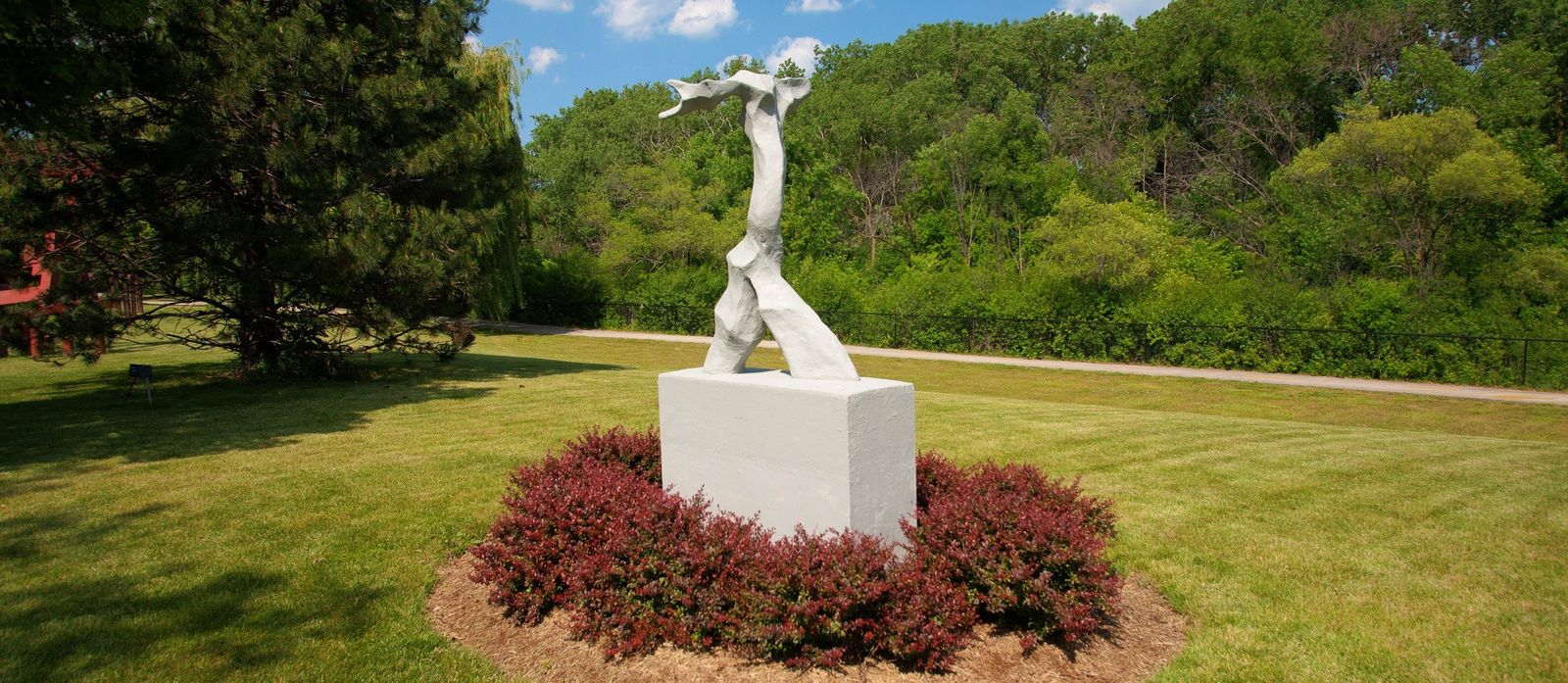 A white sculpture on a plinth ringed by red shrubs on a green lawn with trees behind.