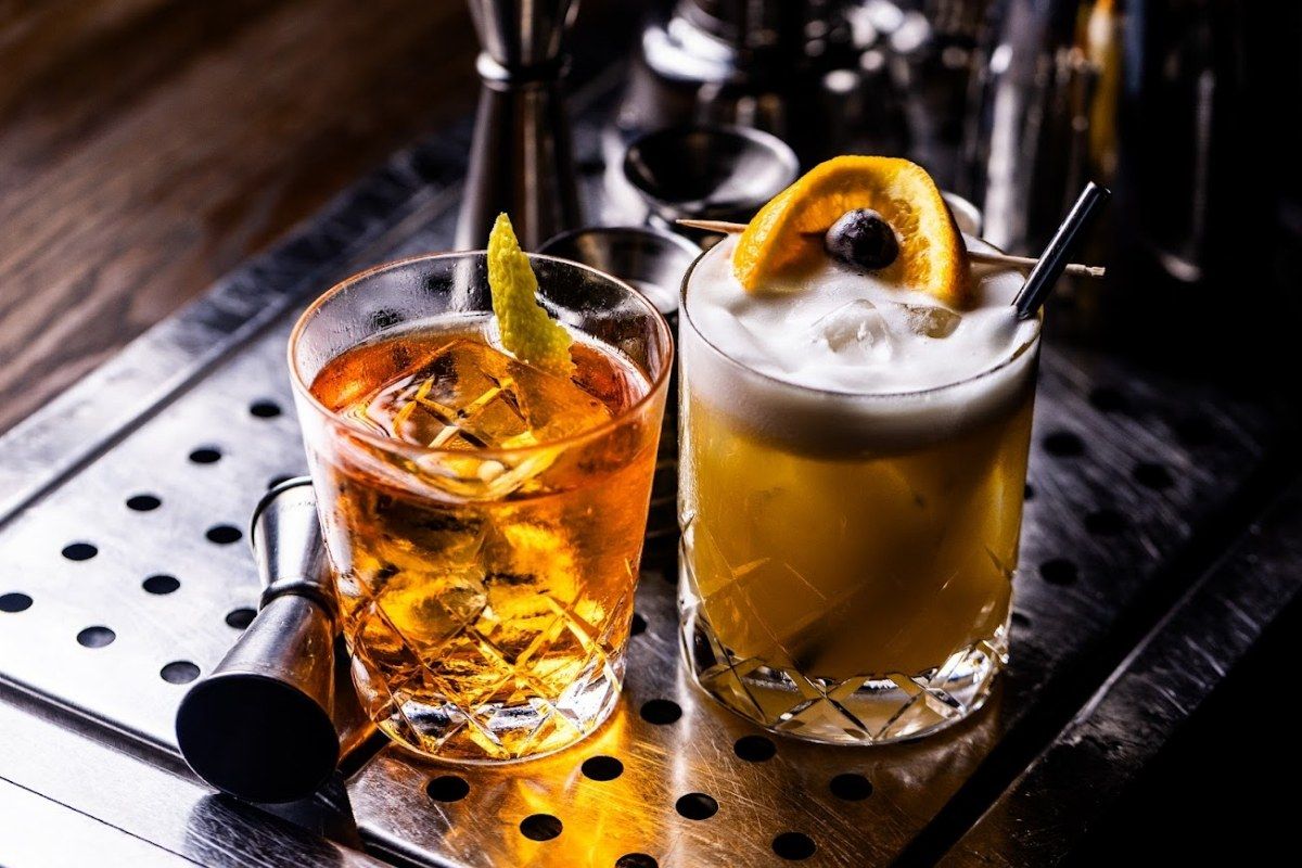Two cocktails garnished with citrus on a perforated metal bar mat with bar tools behind.