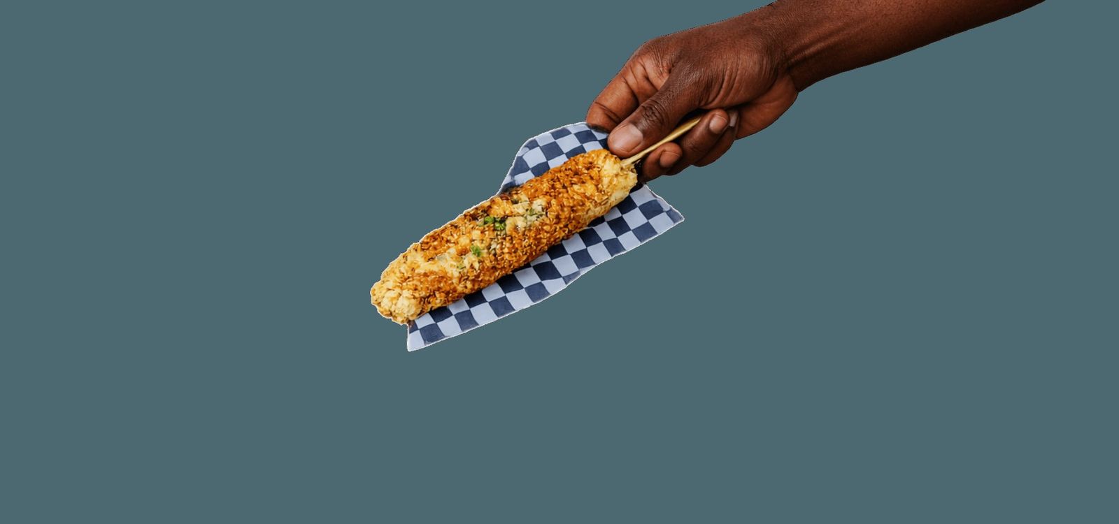 A hand holding a grilled corn on the cob on checkered paper against a gray-blue background