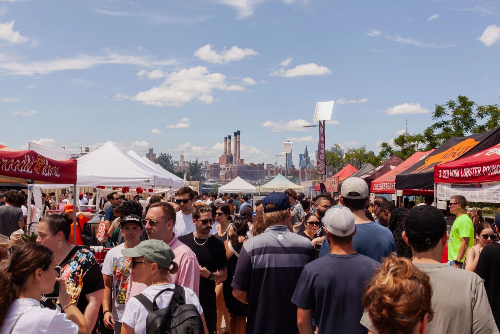 Crowds among market tents under a blue sky with a city skyline beyond the waterfront