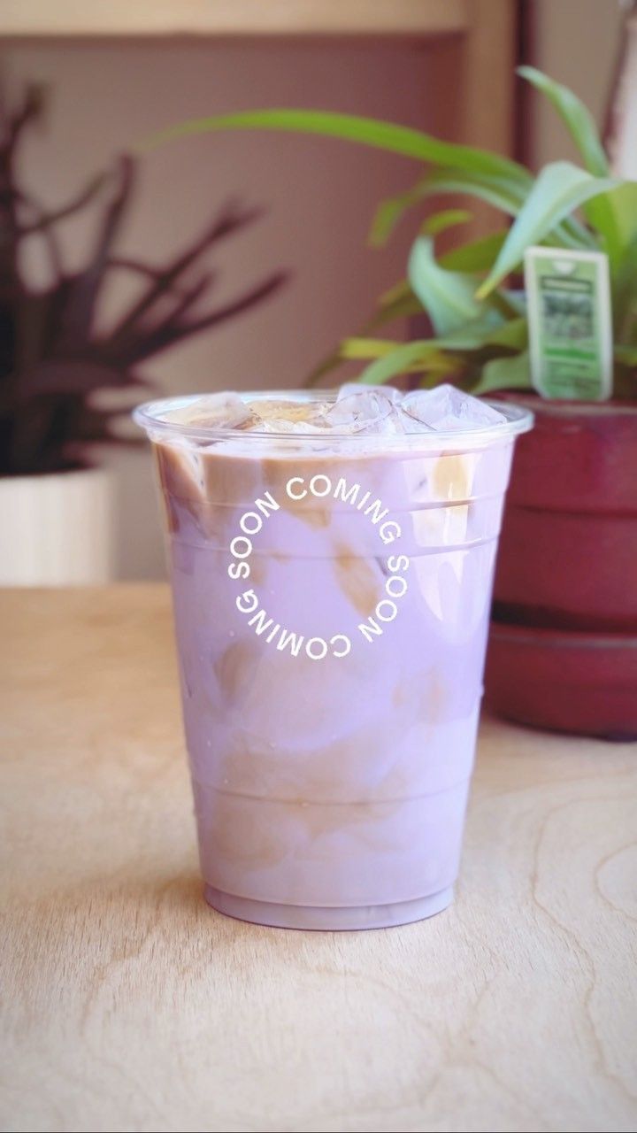 A lavender iced drink in a clear cup on a wooden table, with a plant behind.