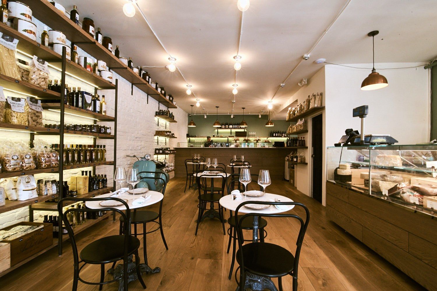 A narrow cafe with small round tables, shelves of goods along the wall and a display counter at the back.