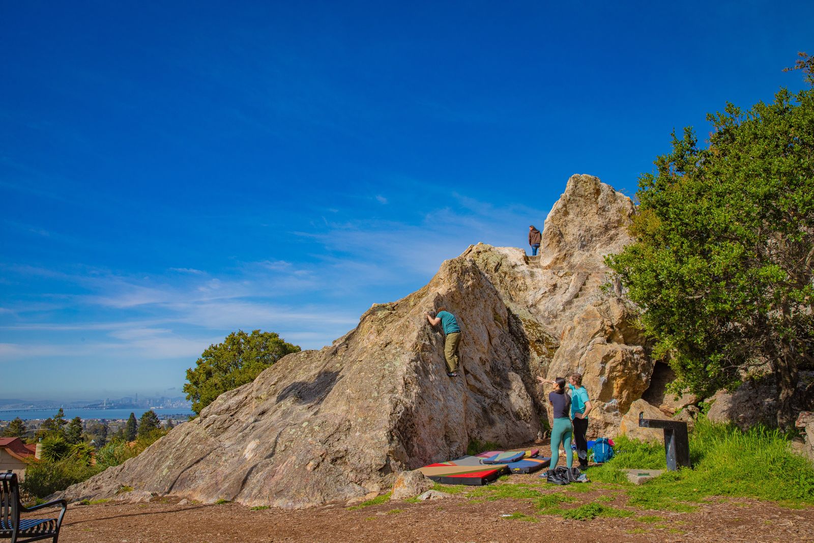 A large granite outcrop with climbers on its face under a blue sky, with grass and trees at its base.