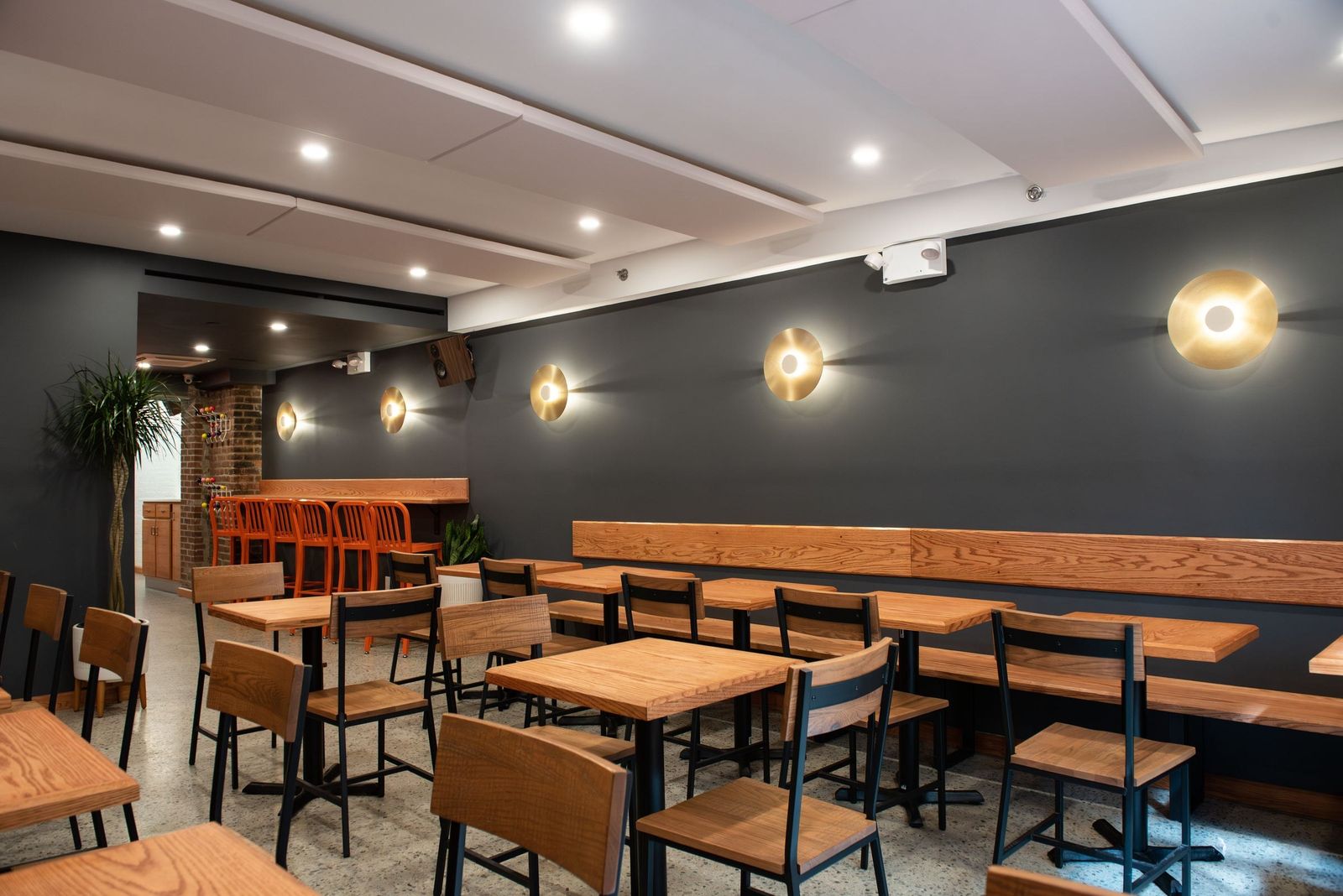 A cafe with wooden tables and chairs, a banquette along a dark wall and round wall lights.