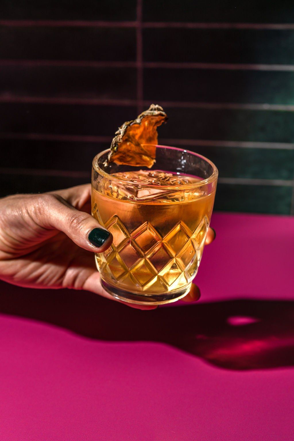 A hand holding a cut-glass tumbler of an amber cocktail with a citrus peel against a magenta background.