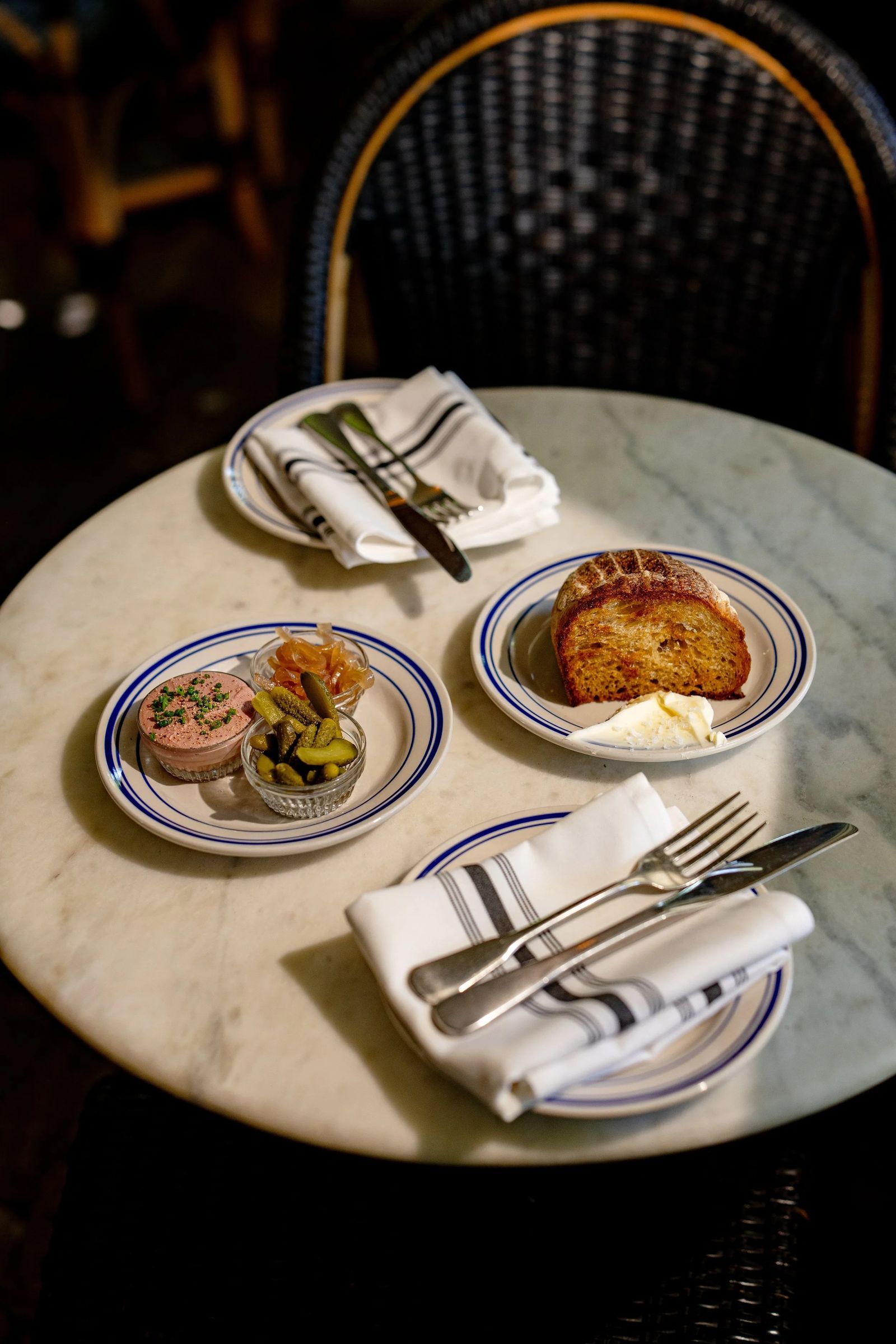 Small plates with blue-rimmed china and folded napkins on a round marble table beside a rattan chair