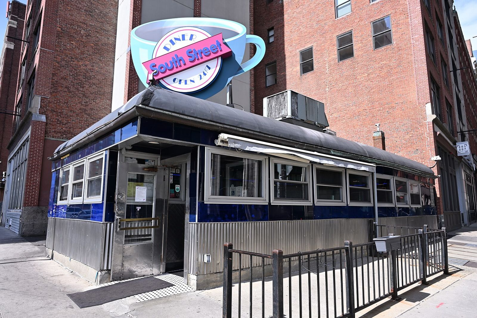 Exterior of a small dark diner building with a coffee cup sign on a brick-lined street