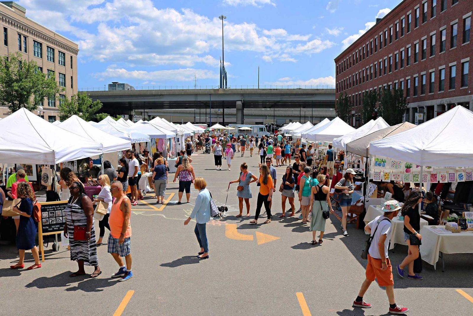 Rows of white tents and crowds along a street at an outdoor market with brick buildings behind
