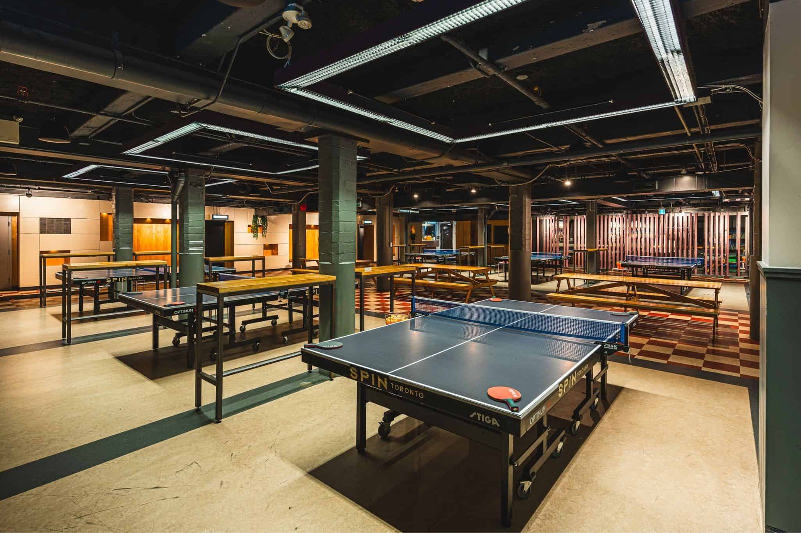 A large room with several ping pong tables under pipes and track lighting, with seating along the walls.