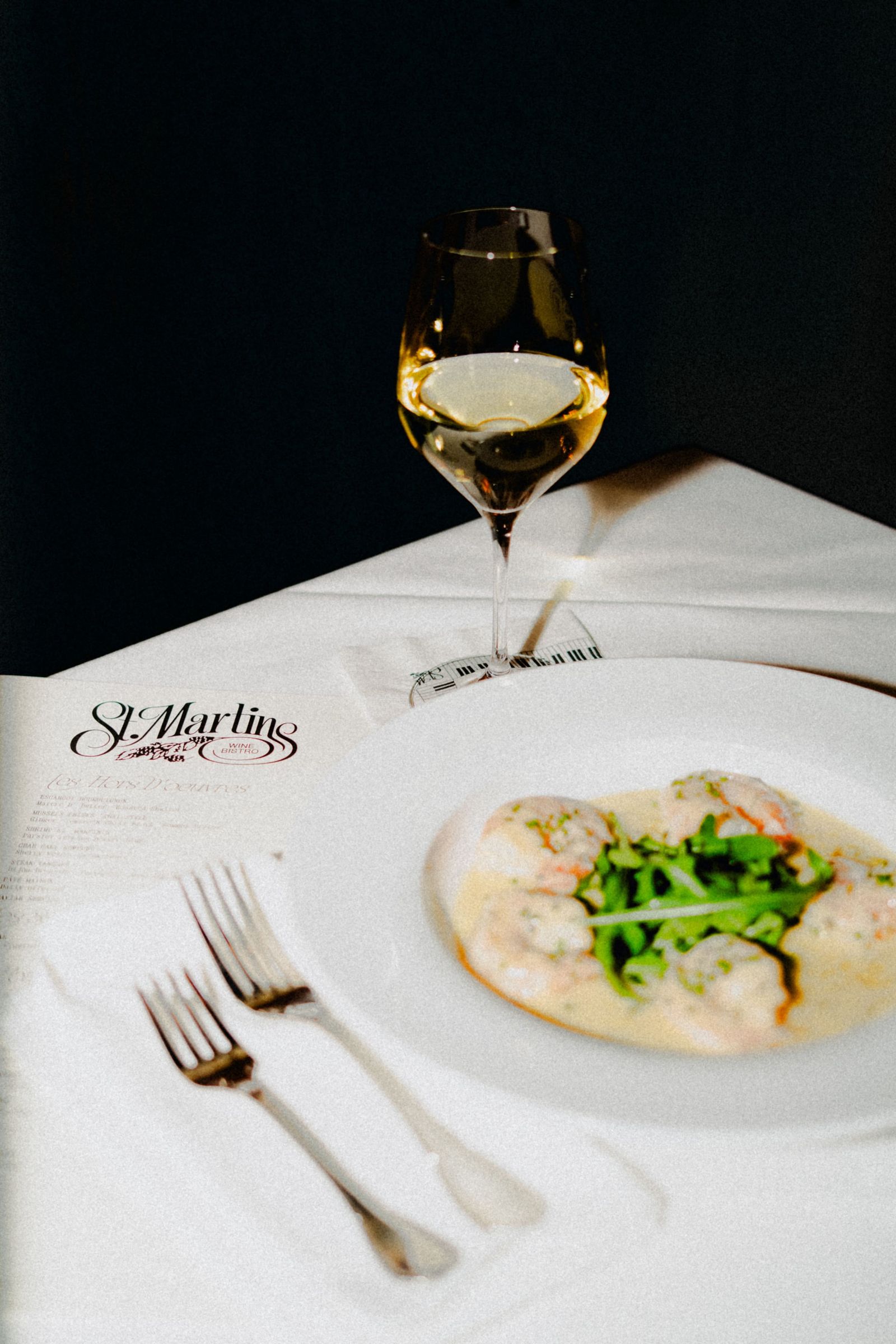 A white plate of pasta with greens beside a glass of white wine on a table with a printed menu.
