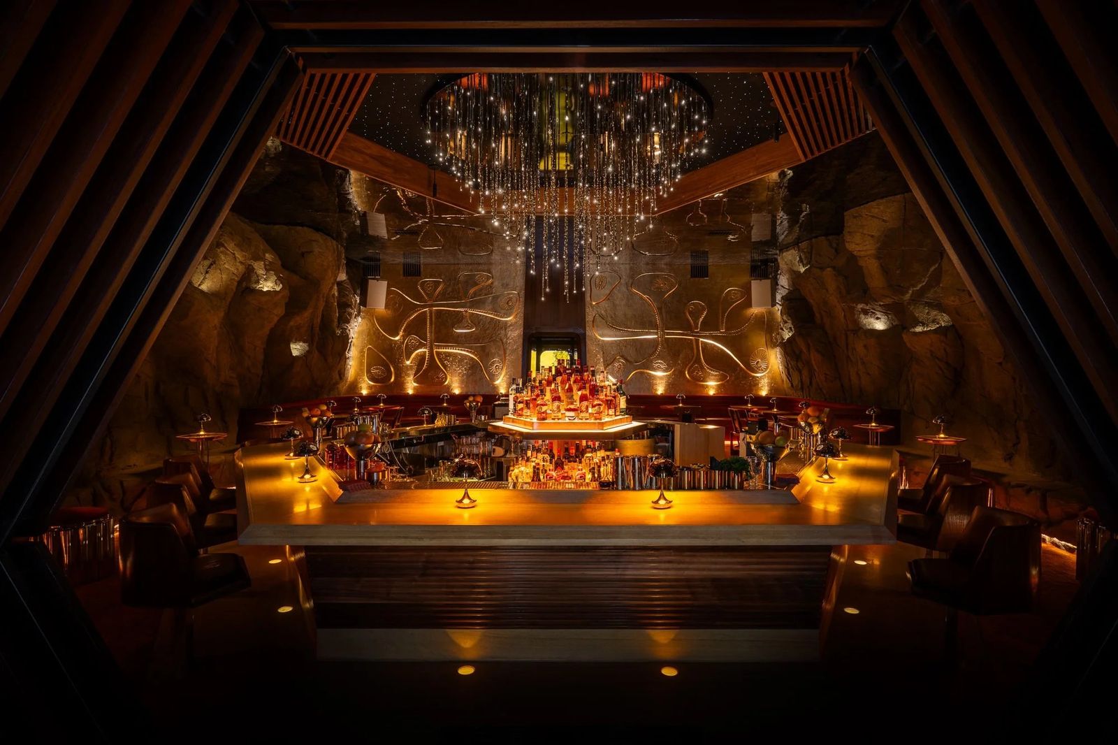 A restaurant lounge with a chandelier, a lit central bar and tables under a dark ceiling.
