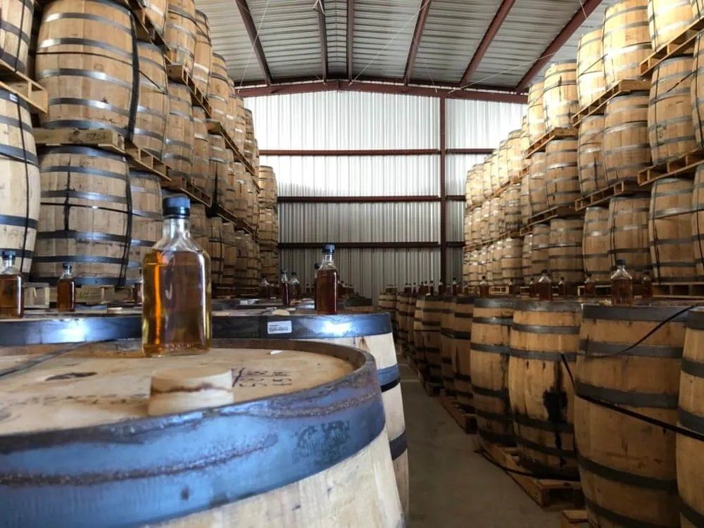 A warehouse with racks of whiskey barrels and a bottle on a barrel in the foreground.