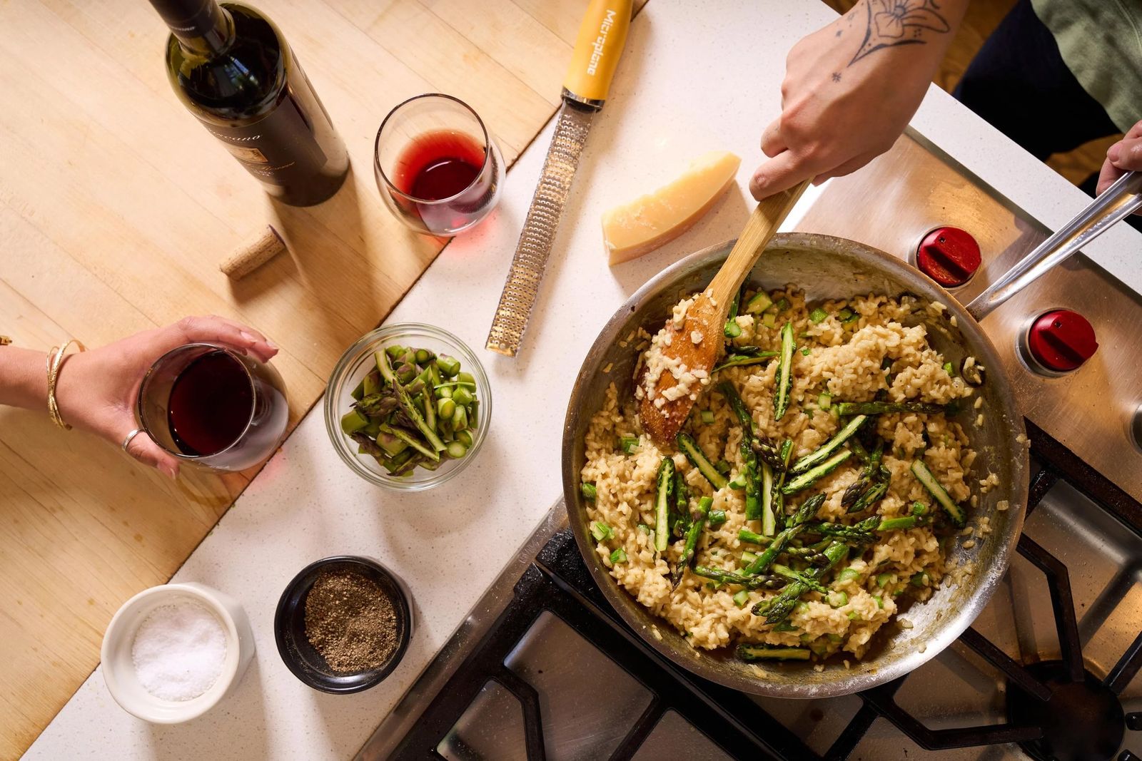 A pan of rice with green vegetables on a gas stove, with a hand stirring and a glass of red wine beside it.
