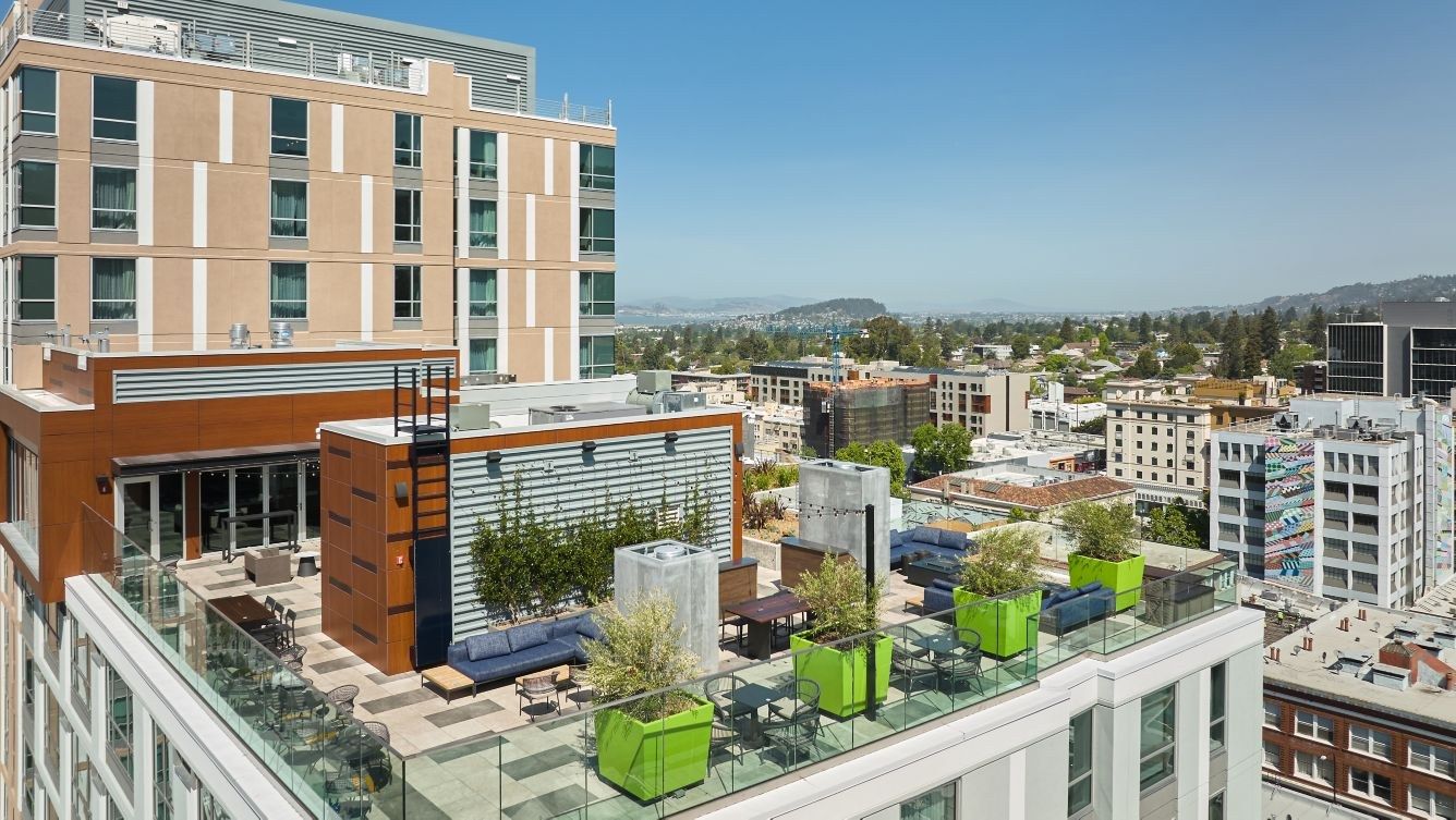 A hotel rooftop terrace with lounge seating and planters above a city street, with hills in the distance.