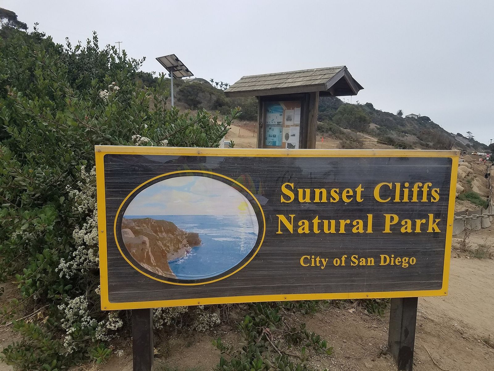 A wooden park sign reading Sunset Cliffs Natural Park, City of San Diego, beside shrubs and a trail board.