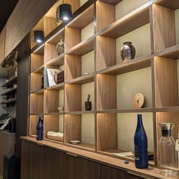 Wooden wall shelving lit from above, holding ceramics and a blue bottle, above a wooden counter with more bottles.