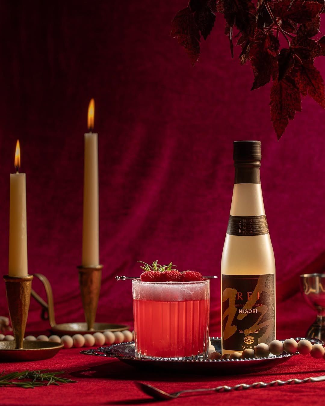 A bottle of sake and a red cocktail on a deep red tablecloth with two lit candles and autumn leaves behind.