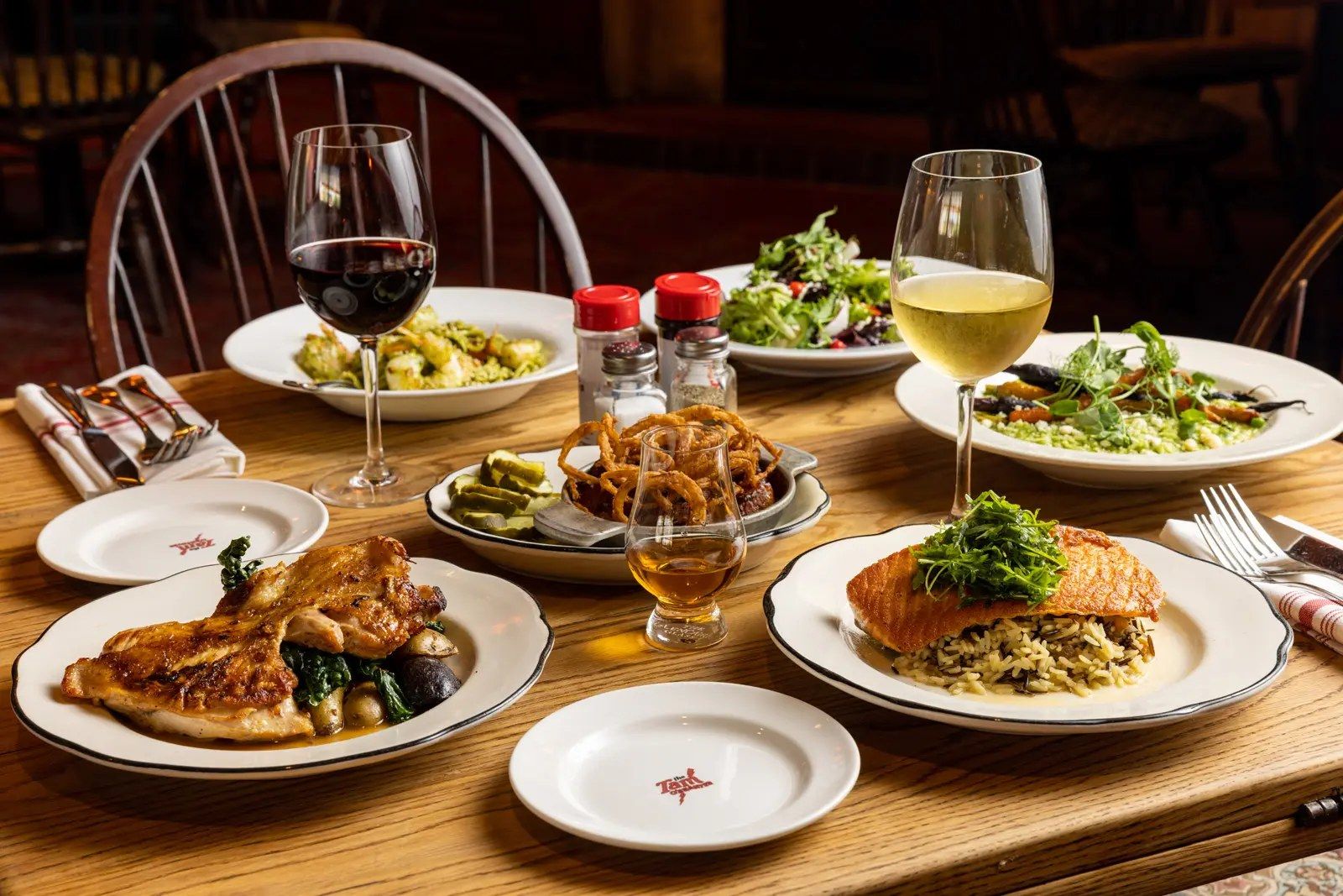 A wooden table set with plates of chicken, salmon and salads, with glasses of red and white wine.