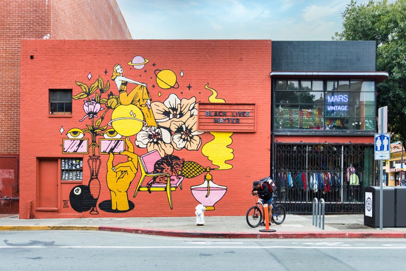 A red brick building with a large painted mural of hands and objects beside a clothing store, with a cyclist passing.