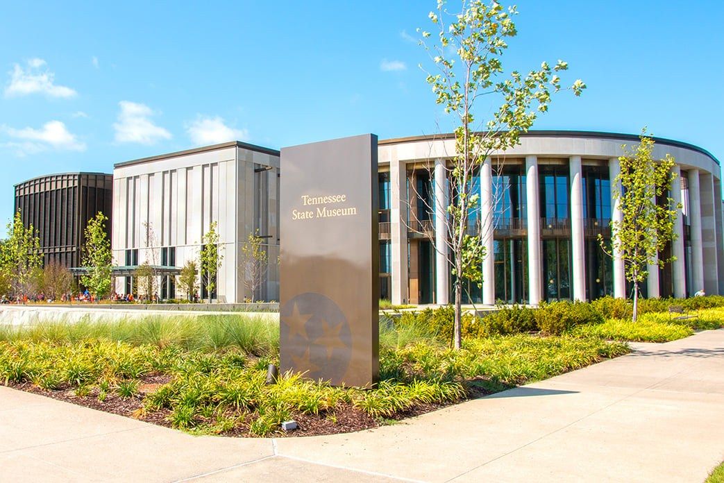 Curved glass-and-stone museum building with a stone sign and landscaped plantings under a blue sky