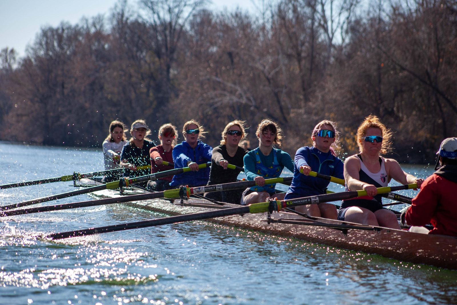 A crew of rowers in an eight-person shell on the river, with trees along the far bank.