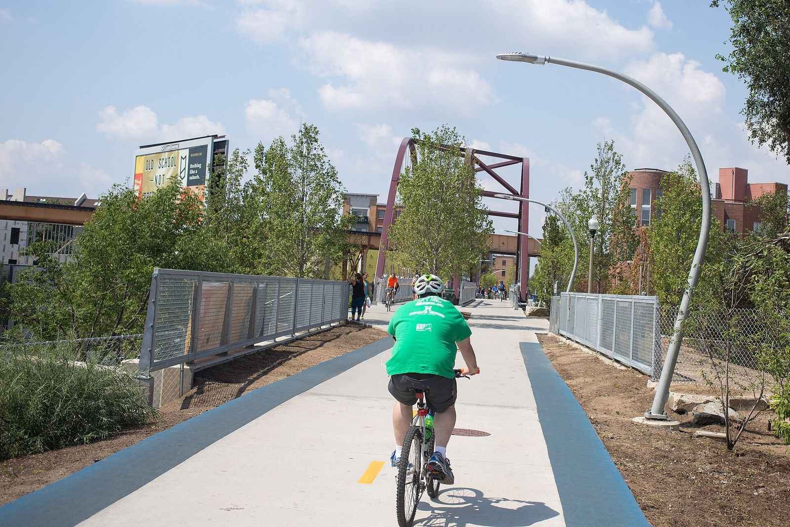 A cyclist in a green shirt on a paved elevated trail with blue edging, with a rust-colored bridge and city buildings behind.