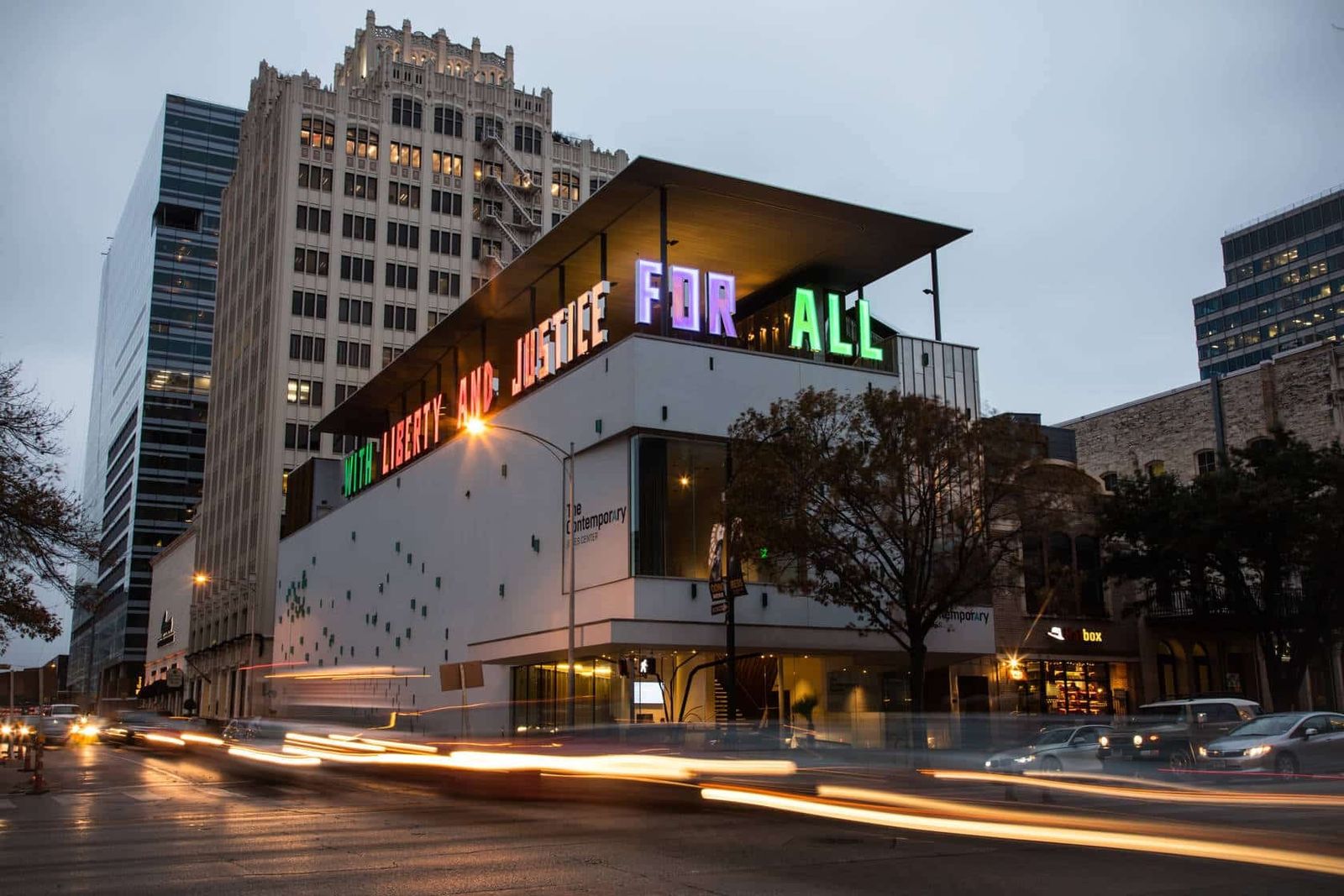The museum building at dusk with a large sign reading For All and the downtown towers behind it.