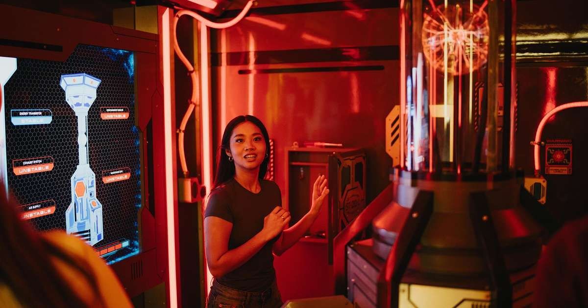 A woman looking at a lit control panel with a glowing screen in a room with red neon lighting.
